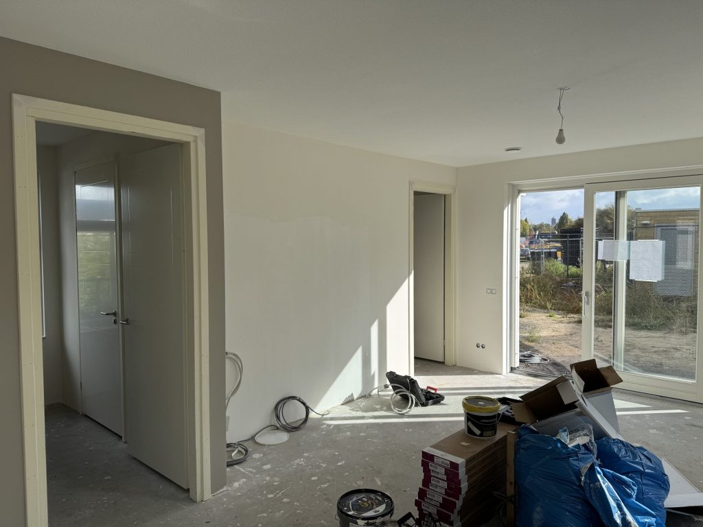 Nieuwbouwwoonkamer tijdens de afwerking, met materiaal op de vloer en een schuifpui naar de tuin