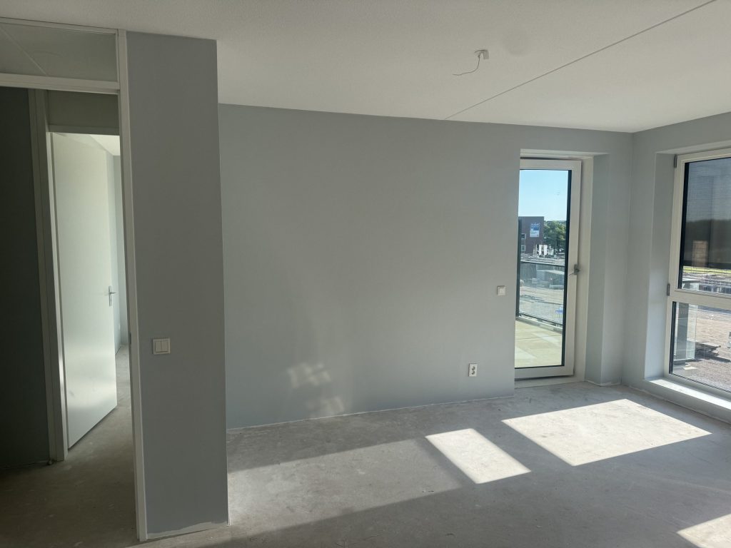 Woonkamer van een nieuw appartement met lichtgrijze wanden, een betonvloer en een balkondeur met zicht op een bouwterrein