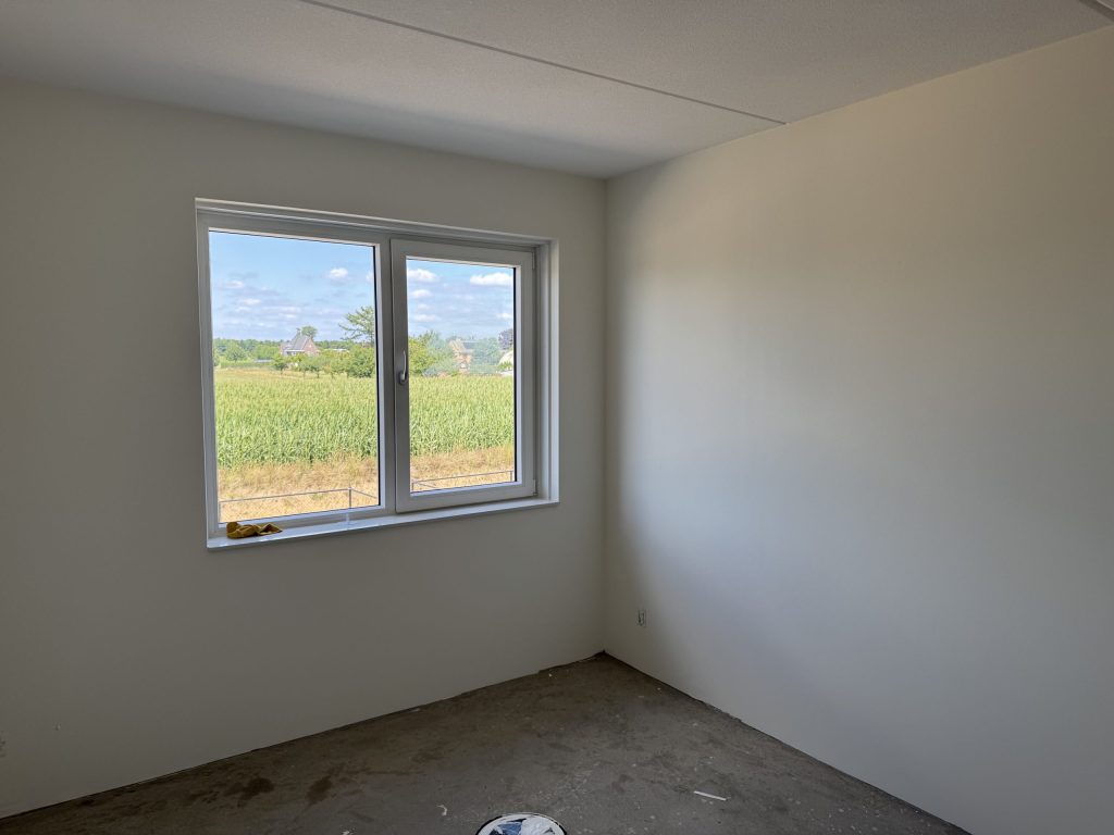 Lege slaapkamer in een nieuwbouwwoning met gladde witte wanden en uitzicht op een maisveld