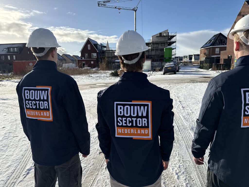 Drie vakmensen van Bouwsector Nederland met witte helm en donkerblauwe jas kijken naar een nieuwbouwwijk met een hijskraan